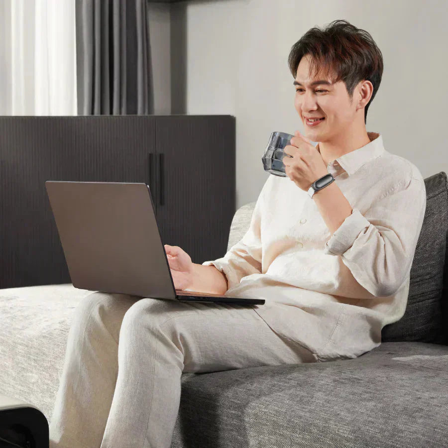 Un uomo sorridente usa un laptop seduto sul divano, indossando un registratore AI indossabile Plaud nero al polso.
