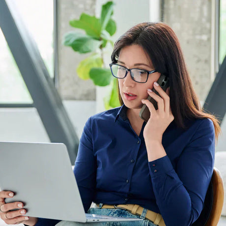 Una donna con occhiali al telefono mentre utilizza un laptop.