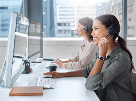 Due donne sorridenti con cuffie lavorano al computer in un ufficio moderno, fornendo supporto clienti.