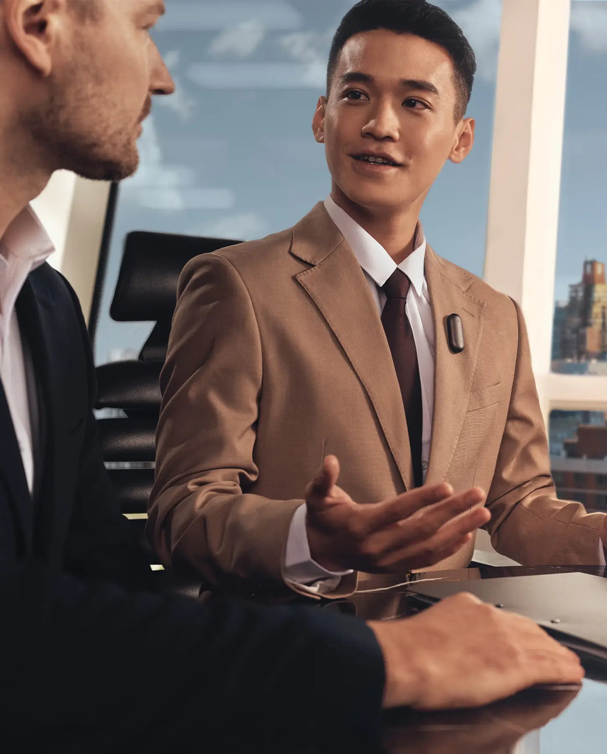 Uomo in abito beige con Plaud NotePin nero sulla giacca, gesticola mentre parla con un collega in un ufficio moderno.